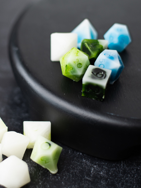 Green, white, blue, and black wax melts shaped like dice on a circular black dish.
